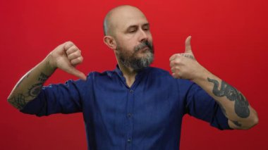 Sakallı kel bir adam, kırmızı arka planda onaylanmış ve onaylanmamış jestler yapıyor, mavi gömlek giyiyor, dövmeler yaptırıyor, yüz ifadelerinde karşıtlık tasvir ediyor..
