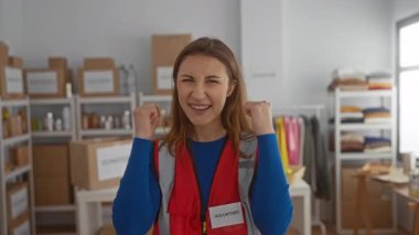 İç mekan hayır kurumunda gönüllü olarak gülümseyen genç bir kadın. Kırmızı yelek giyiyor. Etrafı düzenli kutular, malzeme ve kıyafetlerle dolu..