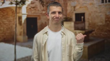 Sıradan kıyafetler giyen bir adam taştan bir arka planı olan bir sokakta dikiliyor, gülümsüyor ve işaret ediyor, güneşli bir günde rahat bir hava veriyor..