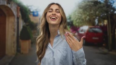 Çizgili tişört giyen sarışın genç bir kadın, güneşli kaldırım taşlı sokakta manikürlü elleri kameraya tutuyor..