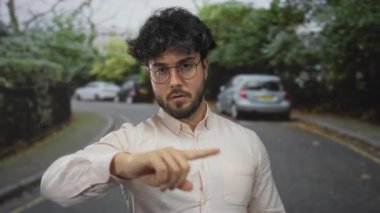 Sakallı genç adam, ağaçlarla çevrili bir sokakta jest yapıyor. Ve gözlüklü ve açık renk gömlekli park edilmiş arabalar.