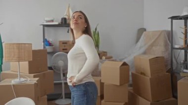Karton kutular ve mobilyalarla çevrili yeni evdeki kutuları açan bir kadın. Parlak bir oturma odasında kolları ve kalçaları üzerinde durarak enerjik bir şekilde gülümsüyor..