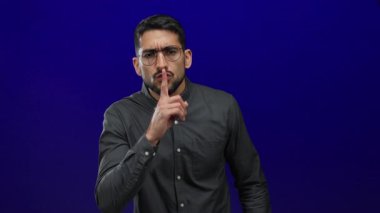 Gözlük takan adam sessiz hareketler yapıyor, parmakları dudakların üzerinde ve yukarı doğru, mavi arka planda izole edilmiş, sessizlik ve dikkat çekiyor..