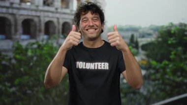 Gönüllü tişörtlü İspanyol bir adam Roma 'nın ikonik kolezyumunun önünde başparmak kaldırıyor, yemyeşil bir zemin ile içten bir gülümseme, pozitifliği ve toplum ruhunu somutlaştırıyor..