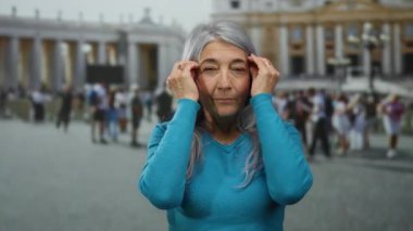 Gri saçlı yaşlı bir kadın canlı mavi süveter giyiyor, Vatikan 'daki hareketli San Pedro Meydanı' nda duruyor, bulanık kalabalık ve tarihi mimariyle çevrili..