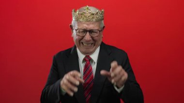 İş elbisesi ve tacı giymiş kıdemli bir adam canlı kırmızı bir arka plana karşı korkmuş ifadeler sergiliyor..