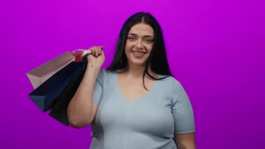 Canlı pembe bir arkaplan önünde elinde renkli alışveriş torbalarıyla gülümseyen bir kadın açık mavi bir gömlek giyiyor ve kendine güvenen bir duruş sergiliyor.