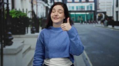 Canlı şehir caddesinde başparmağıyla gülümseyen bir kadın şehir ortamında günlük mavi süveter giyer..