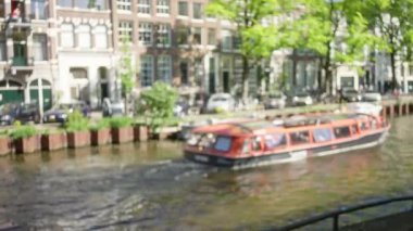 Hollanda, Amsterdam 'daki tekne ve binalar ile bulanık kanal manzarası güneşli bir gün, yemyeşil ağaçlar, park edilmiş arabalar ve kentsel yaşam, sakin bir Avrupa atmosferi yaratıyor.