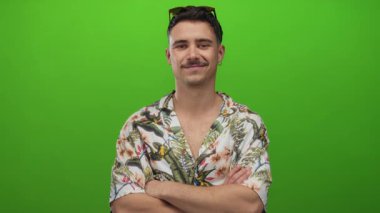 Çapraz kollu İspanyol genç adam çiçekli tişörtle izole edilmiş yeşil arka planda tatilin tadını çıkarıyor, rahat bir turist havası yayıyor..