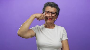 Gri saçlı ve gözlüklü yaşlı bir kadın, parlak bir mor arka plana gülümserken burnunu gösterir, beyaz bir tişört giyer ve neşeli bir jest yapar..