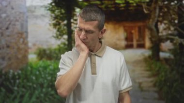 Dışarıdaki taştan yolda duran adam düşünceli ve düşünceli görünüyor. Yeşil yapraklarla çevrili yüzüne dokunuyor..