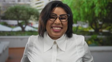 Latin kökenli bir kadın, arka planda canlı yeşillik olan bir parkta neşeyle kaya jesti yapıyor, gözlük takıyor ve beyaz bir gömlek giyiyor, enerjik bir şekilde gülümsüyor..