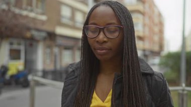 Saçları örgülü ve gözlüklü, şehirde gülümseyen bir kadın, şehir merkezinde ceket giyiyor..