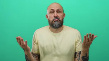 Man in yellow shirt with beard and tattoos shrugging against isolated green background.