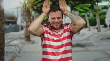 Dövmeleri ve çizgili gömleği olan mutlu bir adam güneşli bir günde arka planda bulanık ağaçlar olan şehir bölgesinde eğlenceli bir şekilde poz veriyor..