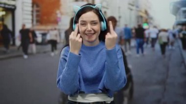 Kadın gülümsüyor, kulaklıkla jest yapıyor, sokak ortamında etrafını sarmış, mavi bir süveter giyiyor, neşeli bir ifade sergiliyor.