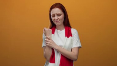 Beyaz tişörtlü, kırmızı saçlı bir kadın turuncu arka planda bileğini acı içinde tutuyor. Eşarp takarken rahatsızlık ve duygu belirtileri gösteriyor..