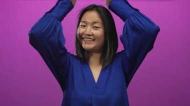 Genç Çinli kadın neşeli bir şekilde gülümsüyor canlı pembe bir arka plana karşı boynuz hareketi yapıyor, neşe ve eğlenceyi ifade ediyor..