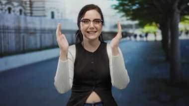 Açık havada esmer bir kadın, sokak sahnesinde ağaçlarla el hareketi yapıyor, gözlük takıyor ve gülümsüyor..