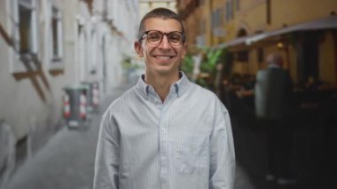 Gözlüklü bir adam Avrupa mimarisi ile çevrili, neşeli bir sokakta duruyor, açık havada neşeli bir anı kucaklıyor..