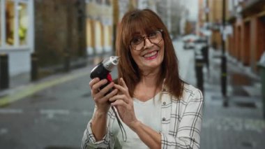 Üst düzey İspanyol kadın, gözlüklü neşe içinde elinde matkapla şehir caddesinde, kareli bir gömlek giyiyor ve arkasında binalar olan parlak bir şekilde gülümsüyor..