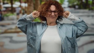 Gürültülü şehir sokaklarında kulakları kulaklarla tıkayan kadın, açık şehir ortamında günlük kot gömlek ve arka planda bulanık elementler içeren gözlük giyerek hayal kırıklığı yaşıyor..