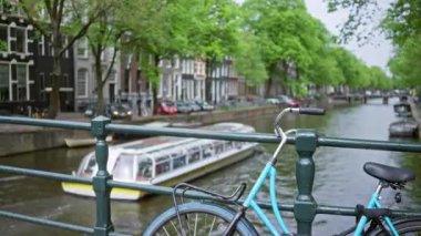 Amsterdam 'da kanal köprüsüne dayanan bisiklet Hollanda' daki su yolu boyunca bulanık tekne ve manzaralı evler bulutlu bir gökyüzü altında kentsel cazibe ve yeşillik sergiliyor..
