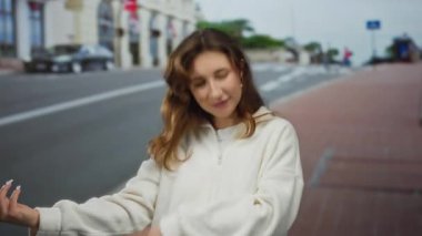 Genç bir kadın şehir sokaklarında bulutlu gökyüzünün altında gelişigüzel beyaz bir kazakla gitar jesti yapıyor. Açık hava ortamında neşe ve yaratıcılığını ifade ediyor..