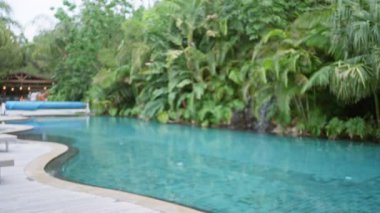 Işıl ışıl parlayan havuzu olan, arka planda yemyeşil ve uzak konuklarla çevrili lüks bir tropikal tatil köyünün bulanık manzarası açık hava ortamının keyfini çıkarıyor..