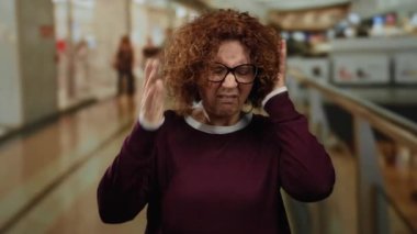 Kıvırcık saçlı bir kadın, kalabalık bir alışveriş merkezinde, yoğun bir alışveriş ortamında stres ve tedirginliğini dile getirirken hayal kırıklığı içinde başını tutuyor..