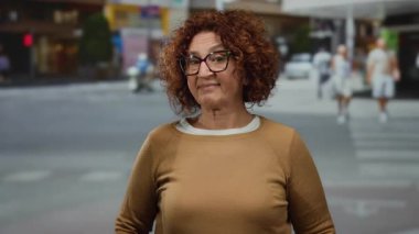 Kıvırcık saçlı ve gözlüklü orta yaşlı kadın hareketli bir şehir caddesinde kendine güvenerek duruyor, şehir hayatını ve farklı kültürleri somutlaştırıyor..