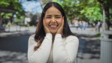 Beyaz kazaklı bir kadın, güneşli bir sokakta neşeli bir şekilde gülümsüyor. Arka planı bulanık insanlar ve yeşil ağaçlarla dolu..