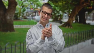 Yeşil ağaçlar ve çitlerle çevrili bir parkta gözlüklü ve gömlekli bir adam doğal bir ortamda mutluluk ve pozitiflik sergiliyor..