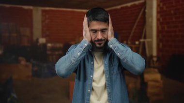 İnşaat alanında kulakları tıkayan genç bir adam, gürültüyle can sıkıntısını ifade eden, sıradan bir kot gömlek ve bej tişört giyen, kapalı arka planda bulanık duran,.