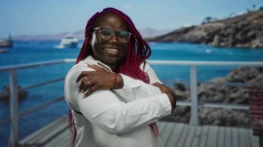 Gözlüklü ve uzun örgülü bir kadın kumsalda neşeyle gülümsüyor, açık bir gökyüzünün altında denizin arka planına ve uzak tepelere sarılıyor..