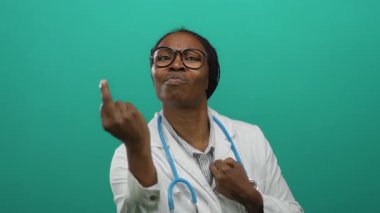 Yeşil arka planda steteskop jestleri yapan, ifadesini ve duygularını gösteren, profesyonel bir tıbbi ortamda çerçevelenmiş, kendine güvenen gözlüklü bir kadın..