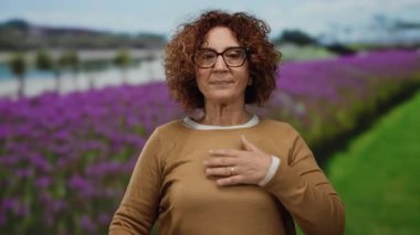 Gözlüklü İspanyol kadın, açık havada renkli bir ortamda kendine güven ve samimiyeti somutlaştıran mor çiçekler ve yeşil tarlalarla dolu canlı bir parkta yemin jesti yapıyor..