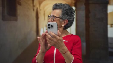 Kırmızı elbiseli, gri saçlı bir kadın kemerli eski bir kasaba caddesinde akıllı telefon kullanıyor. Dijital bağlantı odaklı canlı bir şehir manzarası yakalıyor..
