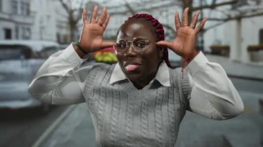 Şehir caddesinde komik suratlar yapan bir kadın, gözlüklü ve gri kazaklı, arka planda ağaçlar ve arabalar olan, açık havada neşeli ve neşeli ifadeler sergiliyor..