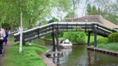 Hollanda, giethoorn 'da bulanık bir kanal sahnesi. Yerinden edilmiş kadın ve adamın yemyeşil ve uzak kayıkçılarla çevrili sakin bir su yolu üzerinde yürüdüğü ahşap bir köprü..