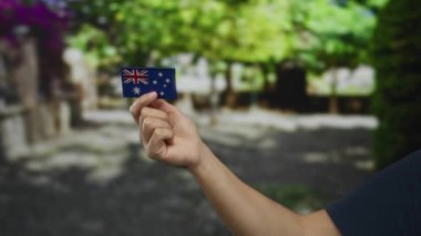 Kafkasyalı bir adam, parkın dışındaki canlı yeşillik ve güneş ışığıyla birlikte açık hava manzarasını aydınlatan Avustralya bayrağını taşıyor..