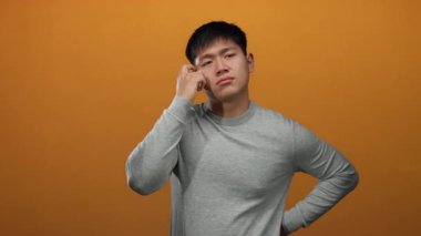 Genç adam turuncu duvara yaslanmış, uykulu görünüyor, gri giyinmiş, yorgun bir yüz ifadesi sergiliyor, parlak bir arkaplanda izole edilmiş, sabah yorgunluğunu ve dinlenmeyi vurguluyor..