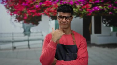 Gözlüklü ve önlüklü genç adam, arka planda çiçek açan canlı bir şehir caddesinde düşünceli bir şekilde el kol hareketi yapar..