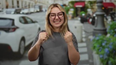 Bir şehir caddesinde yumruklarını sıkarak gülümseyen genç bir kadın, sarı saçlı ve gözlüklü, arka planda arabalar ve çiçeklerle dolu canlı bir şehir ortamında neşe ve başarı çağrıştıran.