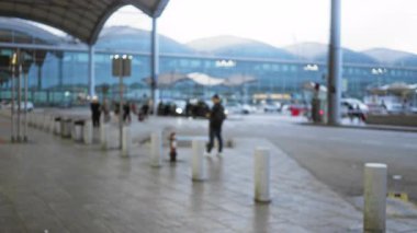 Bulanık görüş, havaalanı terminalinin dışında, bulutlu gökyüzü altında modern mimari ile yürüyen insanların şehirsel seyahat atmosferini arabalarla ve arka planda transit geçişi göstermesi.