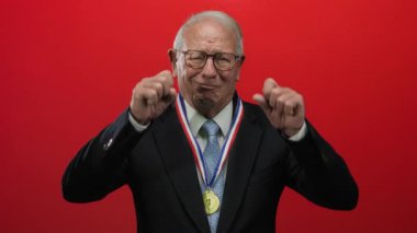 Takım elbiseli kıdemli adam, zafer duygularının mizahi tasviri için parlak kırmızı duvara karşı kazandığı madalyayı takan parmaklarıyla jestleri taklit ediyor..