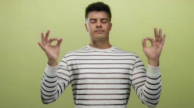 Genç adam, sarı arka planda kapalı gözlerle meditasyon yapıyor, çizgili gömlek giyiyor, sakinlik ve rahatlama taşıyor..