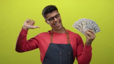 Gözlüklü ve kırmızı tişörtlü, izole edilmiş sarı arka plana gülümseyen, mali başarı ve güven gösteren Amerikan doları tutan bir adam..