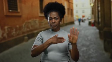 Bir kadın, şehir caddesinde yemin jesti yapıyor, güvenli bir şekilde dışarıda duruyor, tarihi bir kaldırımın altını çiziyor..